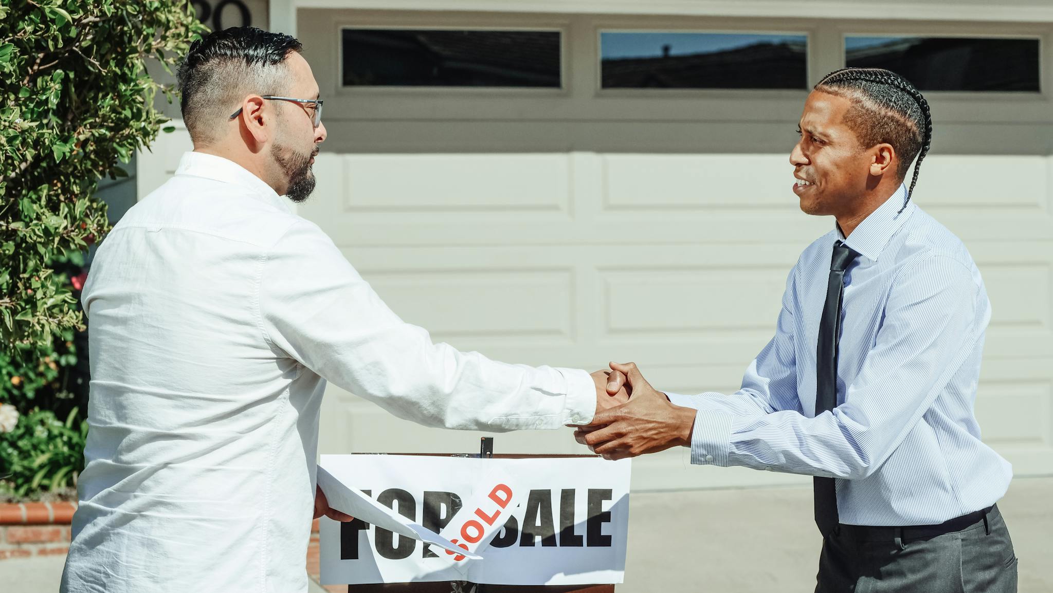 Two men shaking hands in front of a sold house sign, sealing a real estate transaction.
